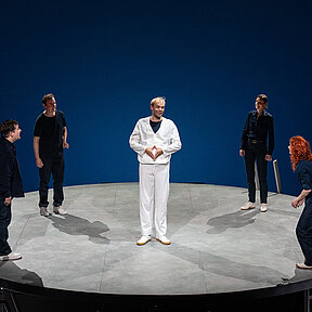 Nico (Hagen Bähr) in weißer Kleidung, er grinst, seine Hände formen eine Raute. Um ihn die anderen Schauspieler:innen.