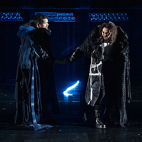 Robert Walton (Paul Enev) und Viktor Frankenstein (Julia Friede) reichen sich die Hand. Düstere Bühnenatmosphäre.