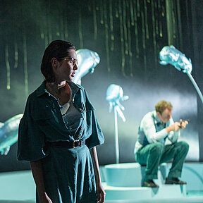 Petra Stockmann (Paulina Hobratschk) steht im Profil, von der Seite beleuchtet. Sie trägt eine Brille und einen grünen Overall. 