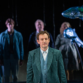 Tomas Stockmann (Klaas Schramm) blickt in die Ferne. Hinter ihm das Bühnenbild und die Stadtgesellschaft sichtbar.