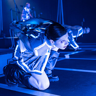 Spieler:innen in gebückter Haltung, sie haben ihre Hände auf dem Rücken verschränkt und nach oben verdreht. Sie gucken gequält, blaues Licht.