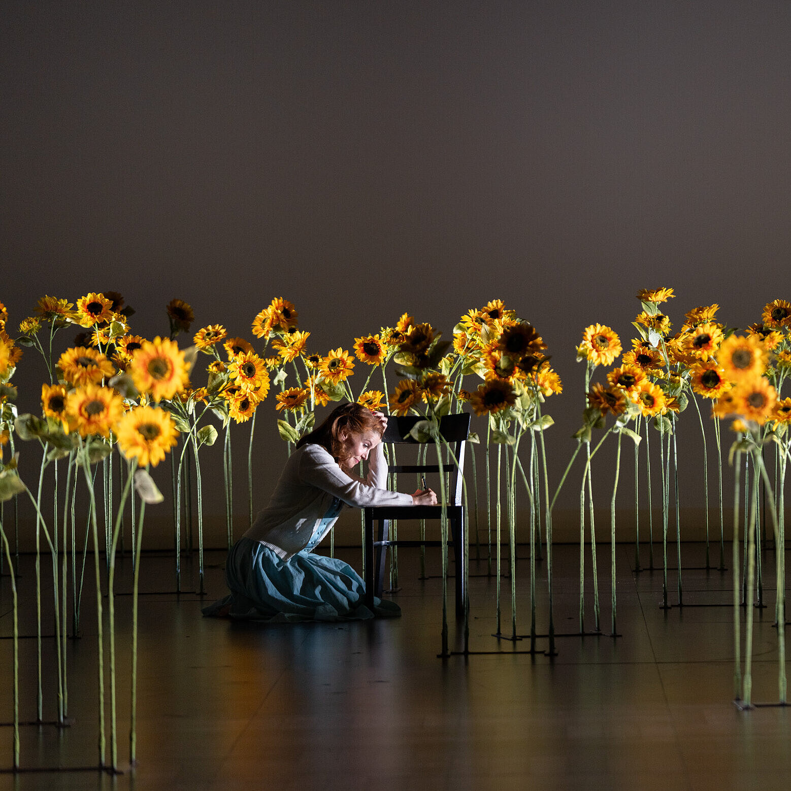 Violetta Valéry (Sarah Vautour) kniet neben einem schwarzen Stuhl in einem Sonnenblumenfeld. Sie schreibt einen Brief.