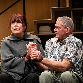 Johanna (Maren Groth-Ische) und Winfried (Benno von Minden) nebeneinander auf dem Sofa. Ihre Hände sind mit einem Gürtel aneinandergefesselt.