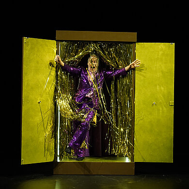 Wolfgang Koch steigt durch einen goldenen Glitzervorhang, der in einem Kasten mit Türen befestigt ist.