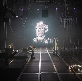 Auf der digitalen Leinwand setzt sich aus weißen Pixeln ein männlicher Kopf zusammen. Die vier stehen verteilt, LED-Streifen leuchten warm-weiß in den dunklen Raum.