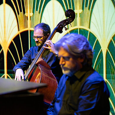 Vor einem grünen Art-Deco Muster sitzen ein Cellist und ein Piano-Spieler