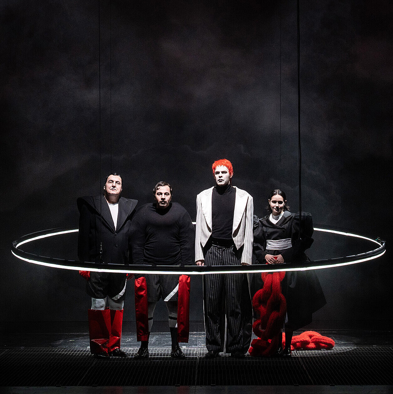 Ensemble steht in auffälligen rot, weiß und schwarzen Kostümen in einem großen Lichtring. 