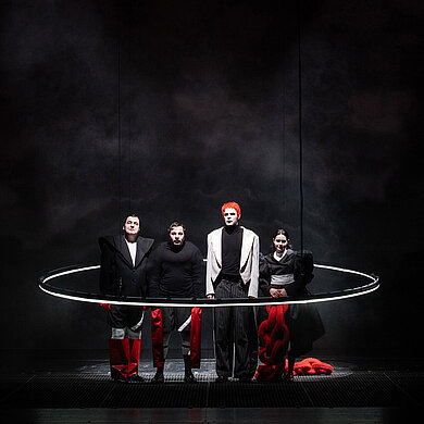 Ensemble steht in auffälligen rot, weiß und schwarzen Kostümen in einem großen Lichtring. 