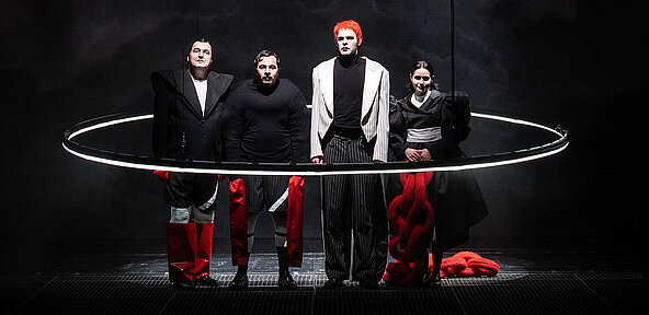 Ensemble steht in auffälligen rot, weiß und schwarzen Kostümen in einem großen Lichtring. 