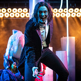 Viktor Frankenstein (Julia Friede) mit einer Überlegenheitsgeste. Im Hintergrund runde Scheinwerfer und halbtransparente aufrechtstehende Rollen.