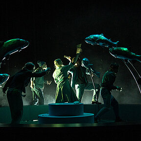 Fünf nur schemenhaft erkennbare Personen, die in einem Brunnen stehen und kleine Fische in eine Richtung werfen. Wenig Beleuchtung, grün-blauer Schimmer. 