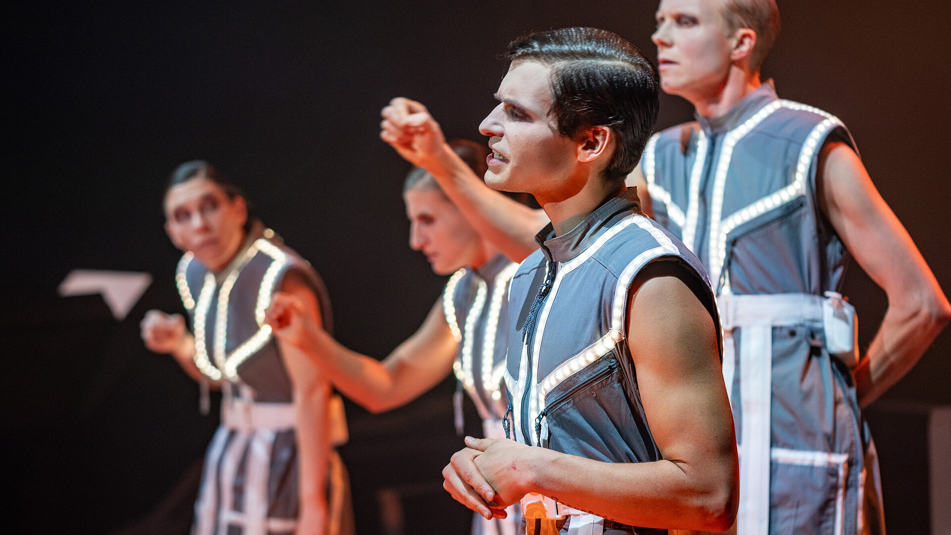 Konstantin Gries vorne im Seitenprofil. Unscharf hinter ihm stehen weitere Spielende schreiben pantomimisch in der Luft. Alle tragen graue schulterfreie Kittel, auf die leuchtende LED-Streifen genäht sind.