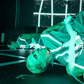 Tobias Schormann liegt zusammengekauert auf dem Boden, mit dem Blick zur Decke. Im Hintergrund liegt Konstantin Gries, in leidender Haltung.